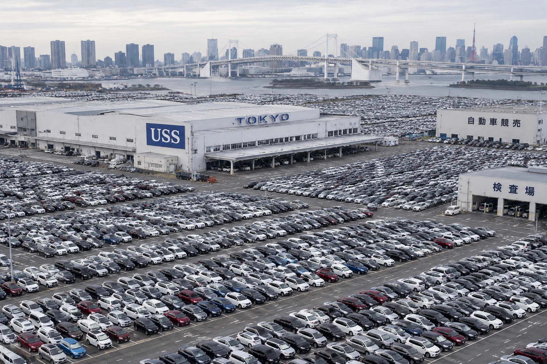 Illustrative straight-down aerial view of a vast Japanese auction yard with dense rows of parked vehicles and a USS Tokyo facility building